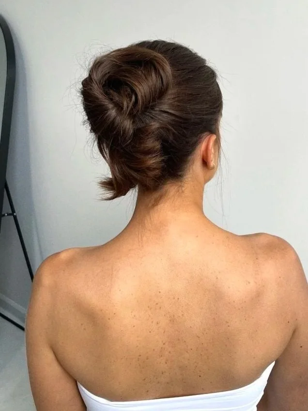 Back view of a woman with brown hair styled in an elegant bun, wearing a white strapless top, showing her shoulders and back.