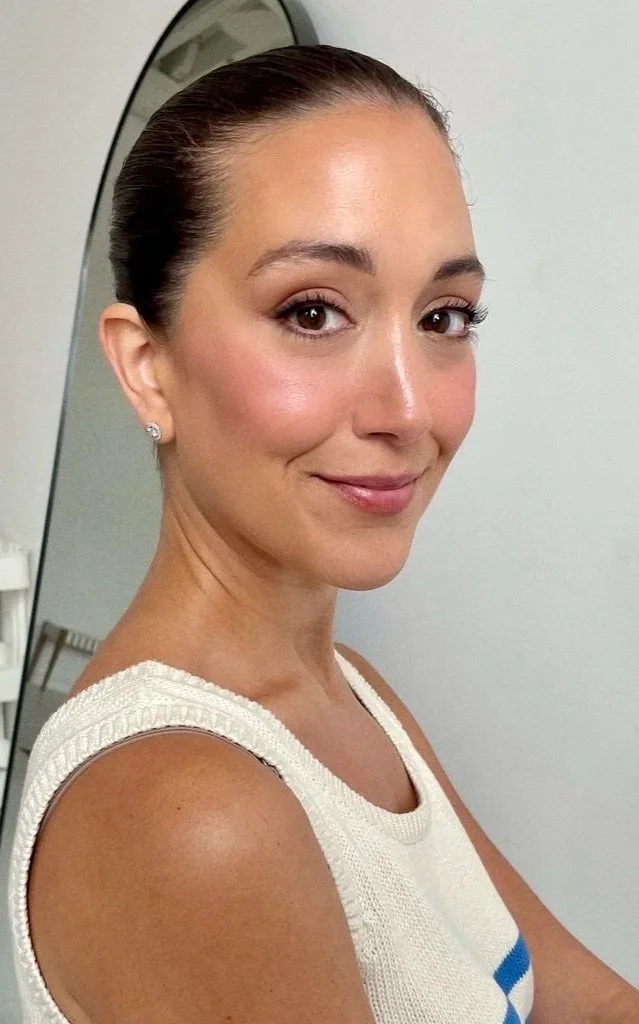 Close-up of a woman with a neat, slicked-back hairstyle, wearing pearl earrings, a white sleeveless top, and smiling softly, standing in front of a mirror.