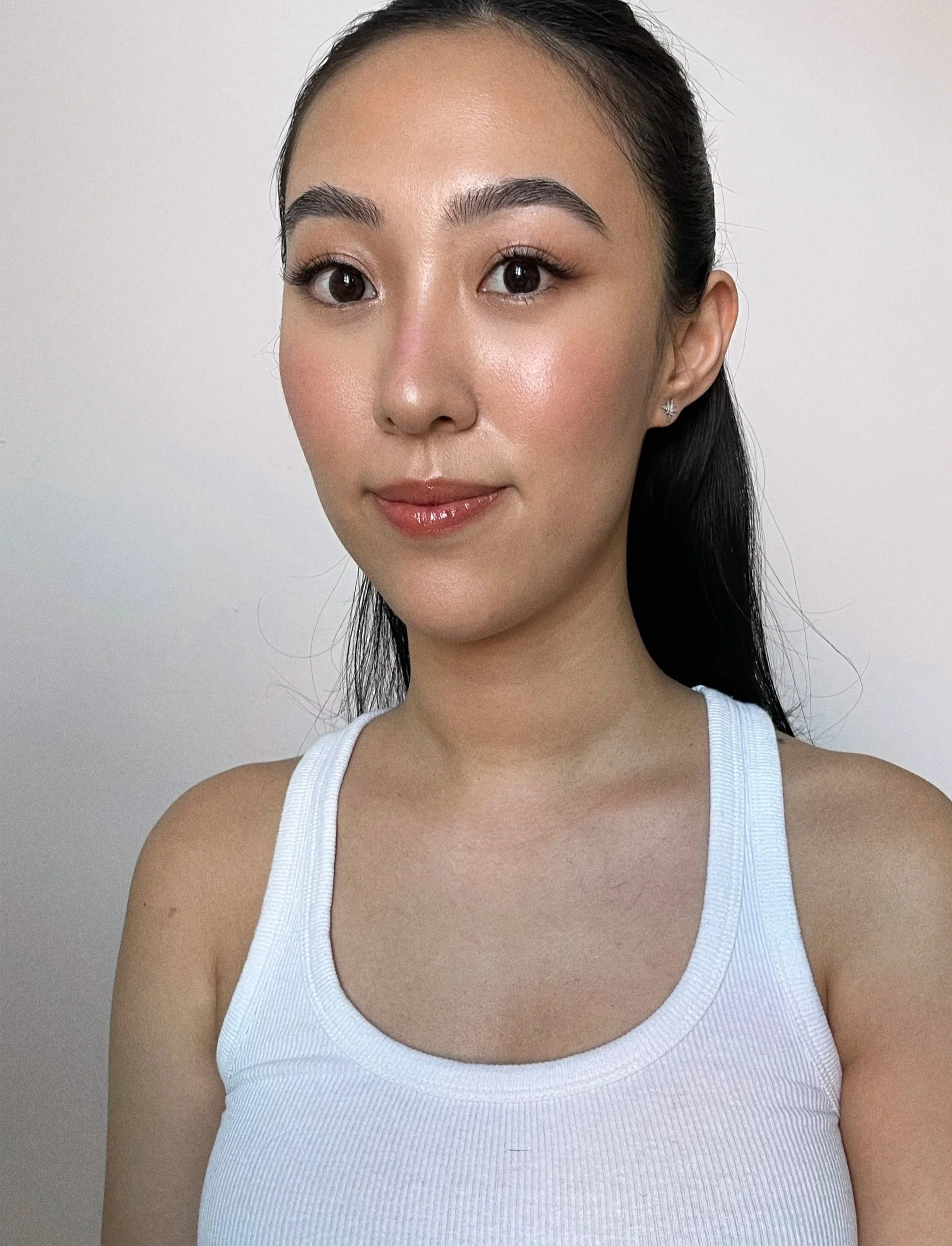 A young woman with fair skin, dark hair tied back, wearing a white tank top, and small earrings, looking at the camera against a plain background.