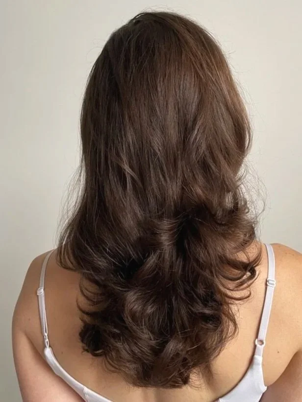 Back view of a woman with shoulder-length, wavy brown hair wearing a white top with thin straps.