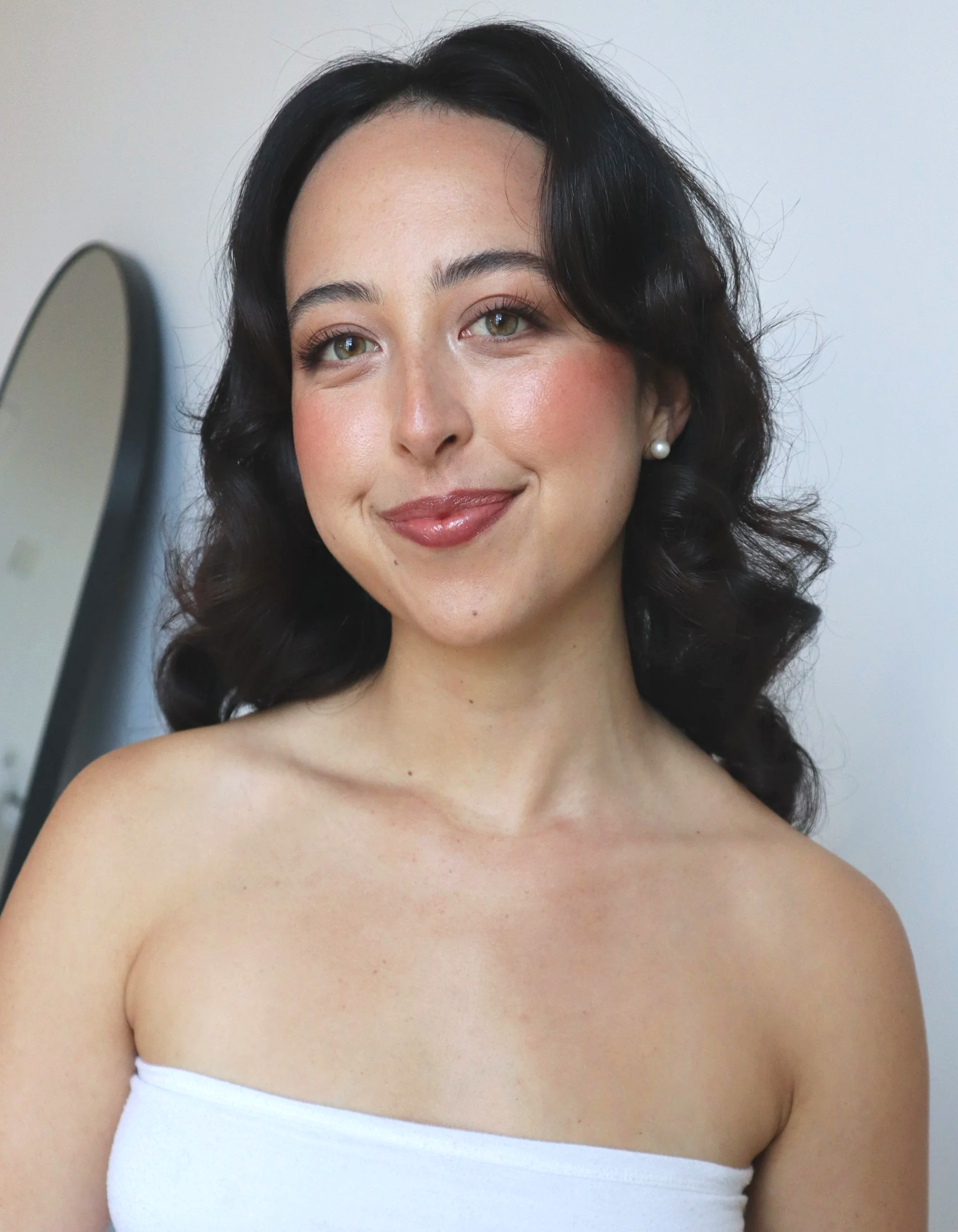 A young woman with dark, wavy hair and light skin, smiling softly and looking directly at the camera. She is wearing pearl earrings and a white strapless top, standing in front of a plain wall with a mirror in the background.