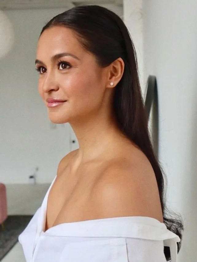 A woman with long, dark hair and earrings, wearing an off-the-shoulder white top, looking to the side in a bright indoor setting.