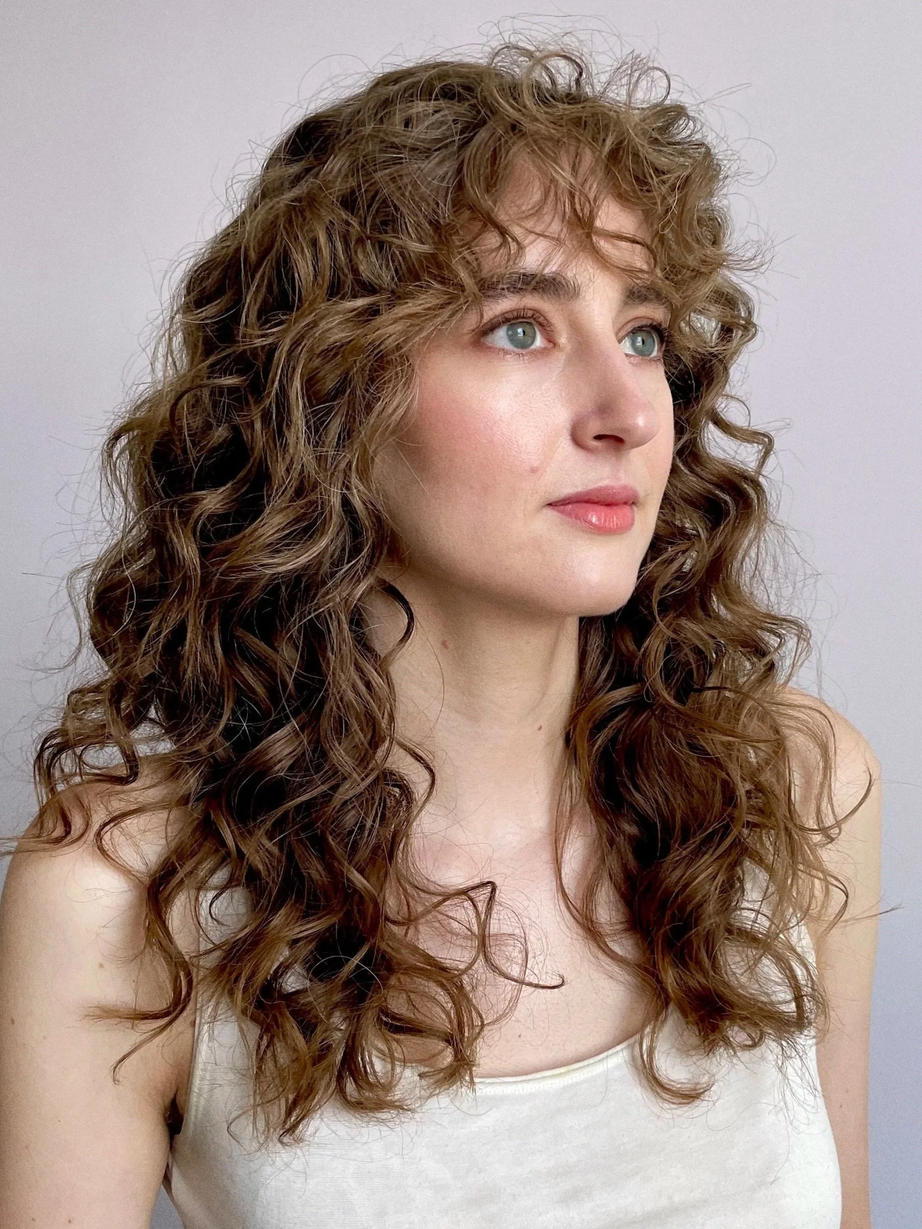 A woman with curly, light brown hair and blue eyes gazing slightly to the side against a plain light gray background.