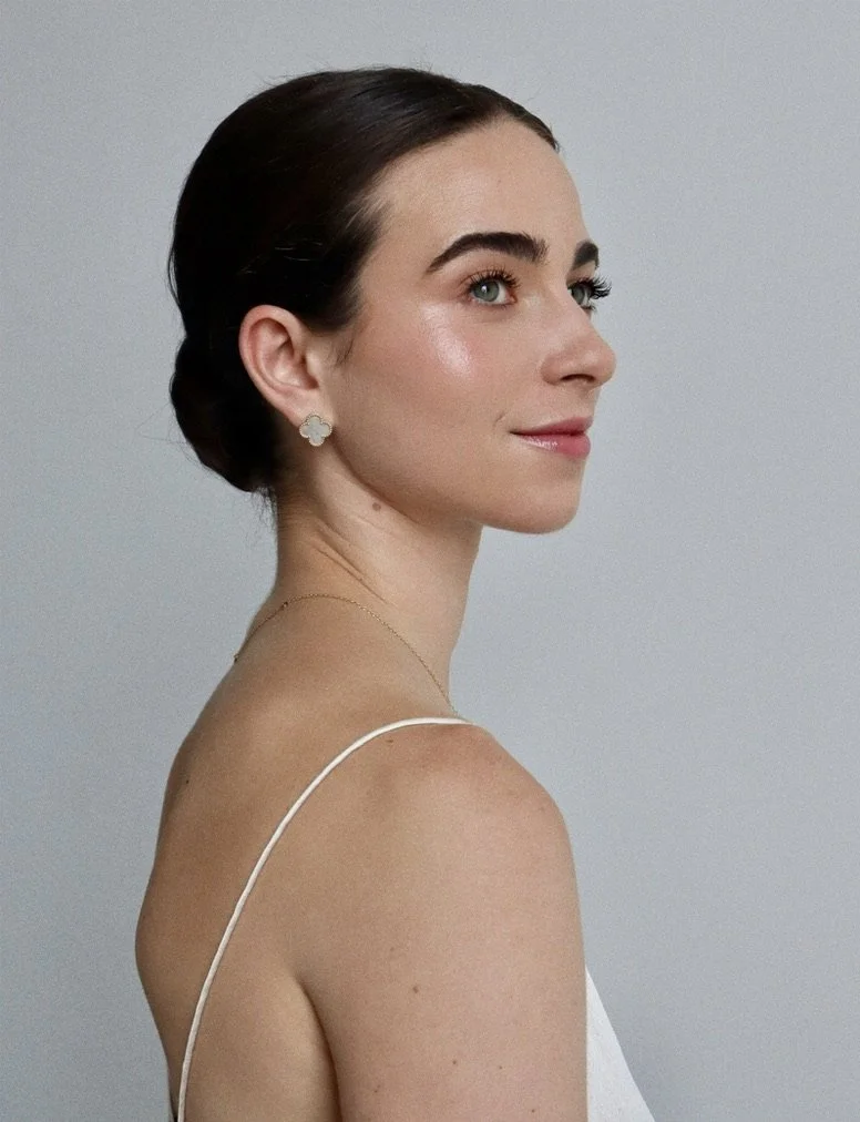 A woman with dark hair styled in an elegant updo, wearing flower-shaped earrings and a thin gold necklace, looking to the side with a subtle smile, against a plain light gray background.