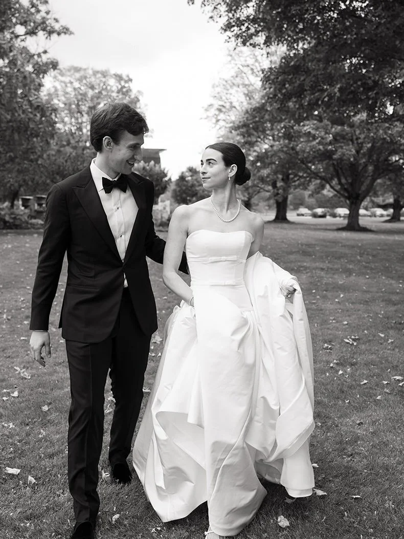 A black and white photo of a bride and groom outdoors. The groom is wearing a black tuxedo with a bow tie, and the bride is in a strapless white wedding dress, holding part of her dress with her right hand. They are smiling at each other, standing on
