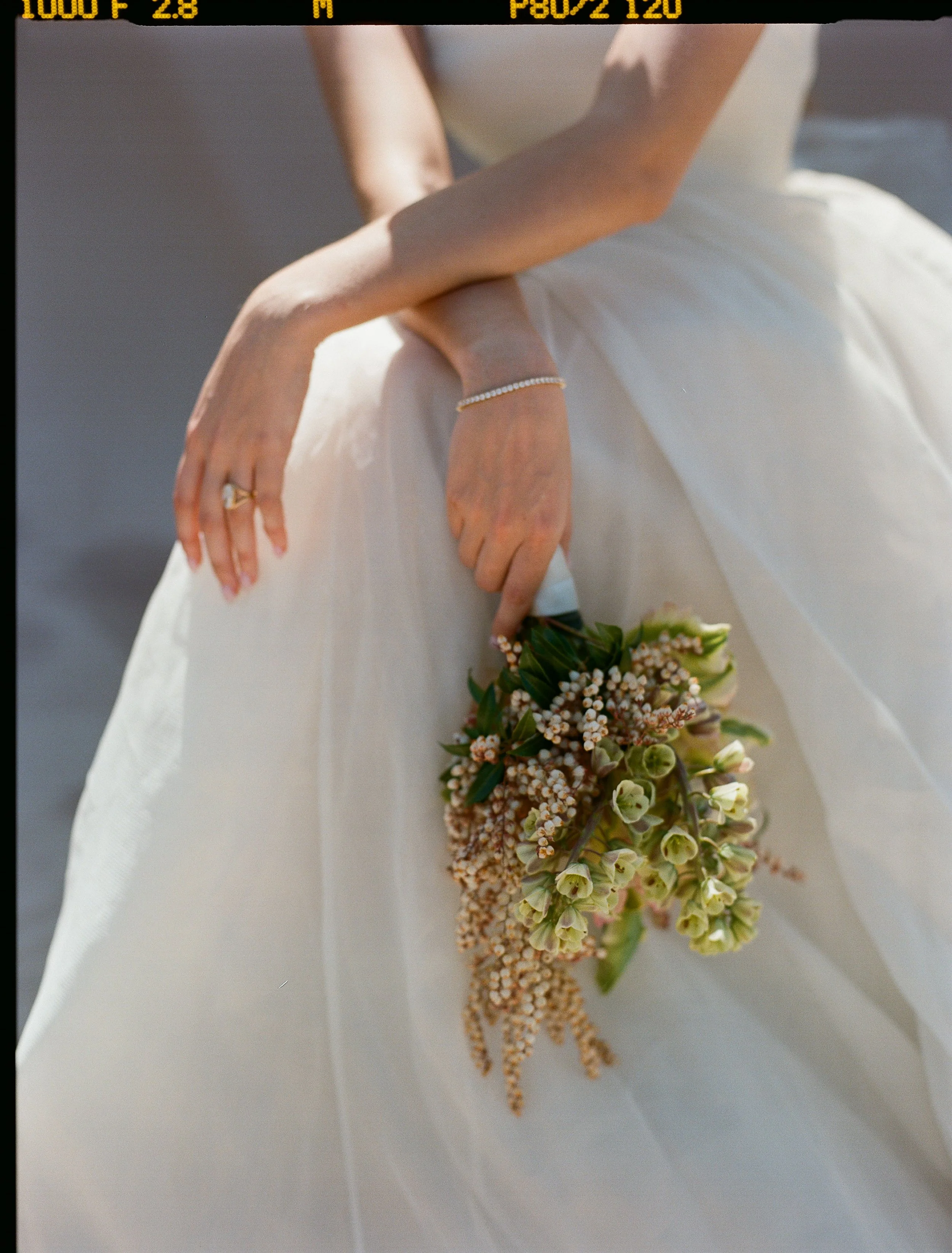 A bride in a white wedding dress holding a cascading bouquet of flowers, with her left hand resting on her skirt, wearing a pearl bracelet and a ring.