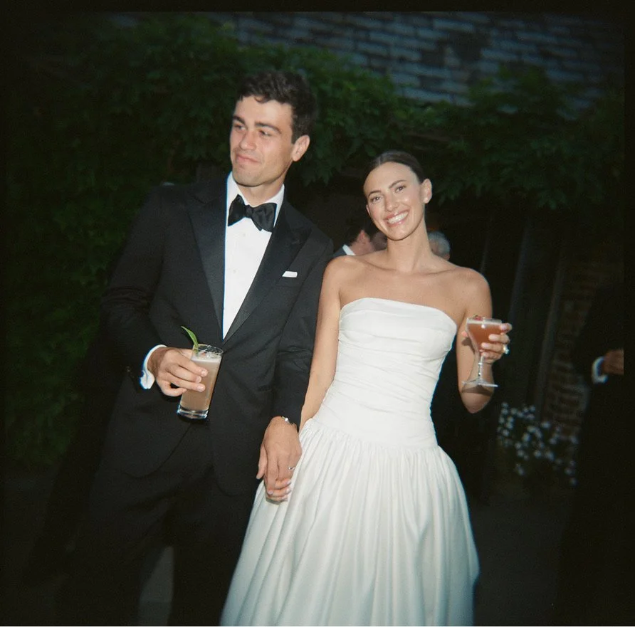A couple dressed in formal wedding attire, holding drinks, at a celebration or wedding reception.