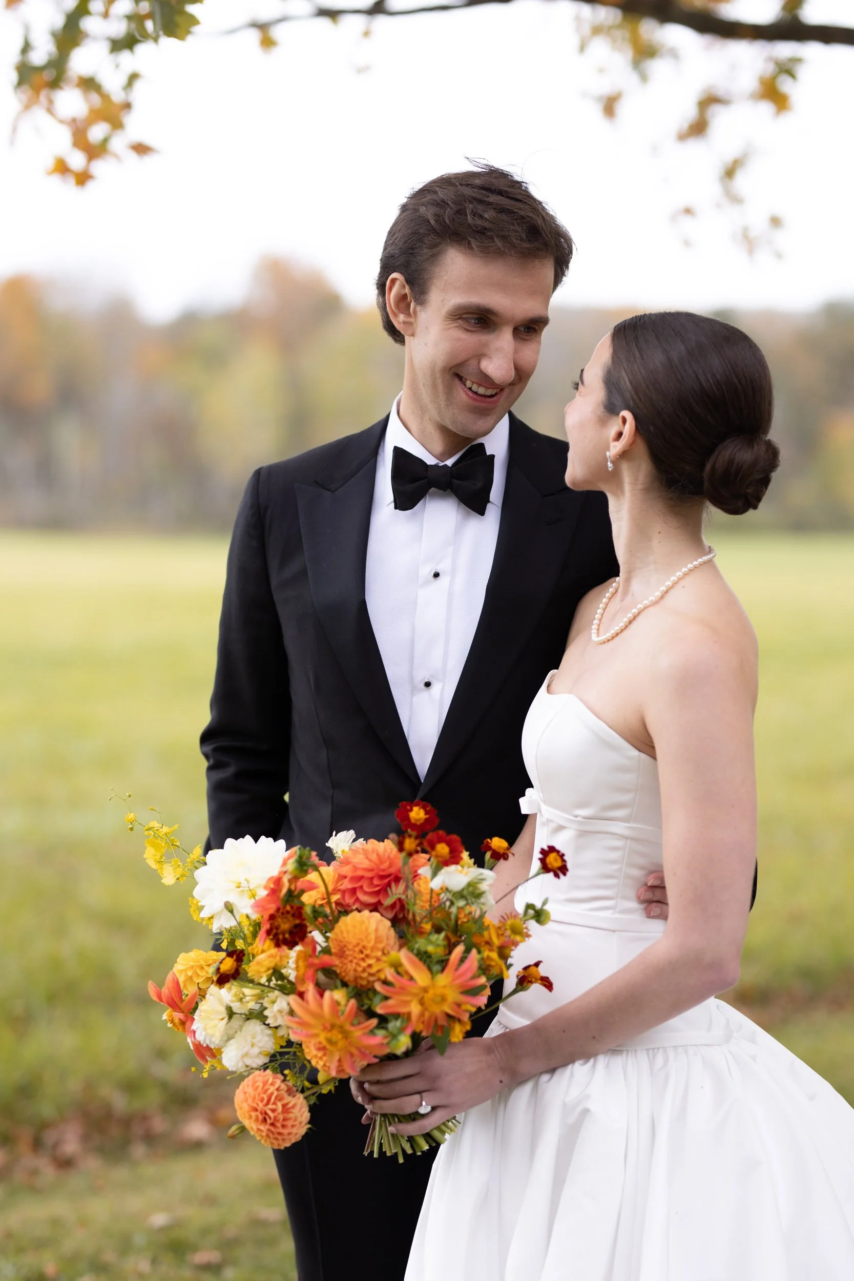 A bride and groom dressed in wedding attire standing outdoors, the bride holding a colorful bouquet, smiling at each other with autumn trees in the background.