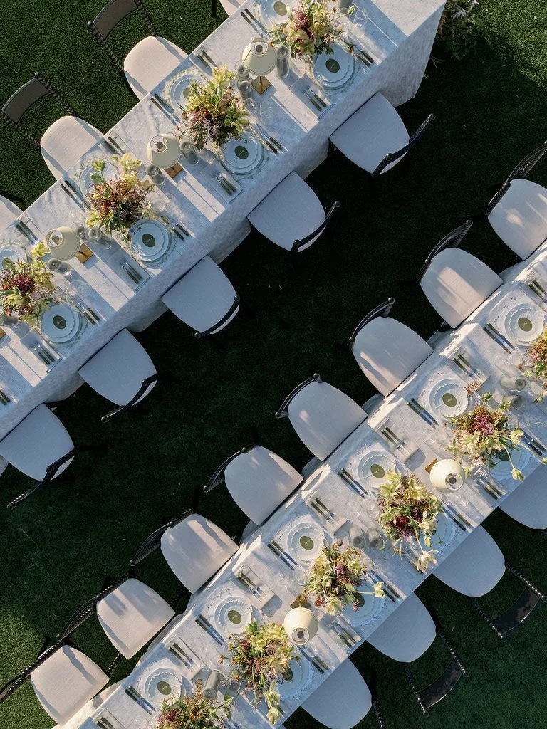 Overhead view of a long outdoor dining table set for a formal event with white tablecloths, floral centerpieces, plates, silverware, and glasses, surrounded by white and black chairs on a grassy area.