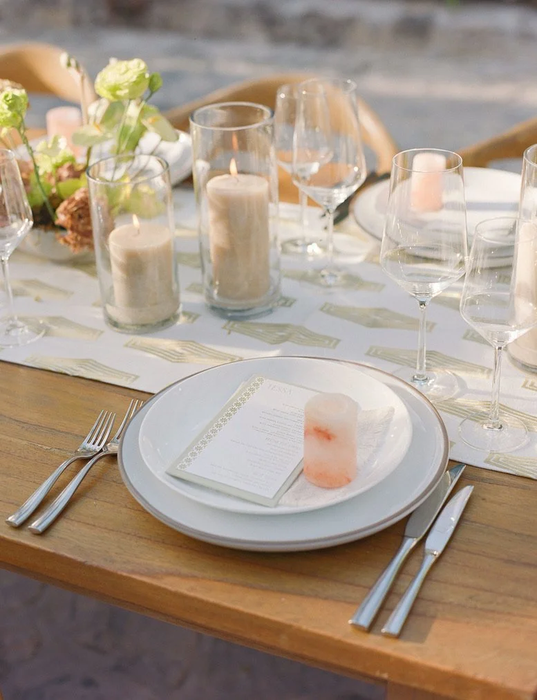 Elegant table setting with white plates, silverware, wine glasses, candles in glass holders, and a floral centerpiece.