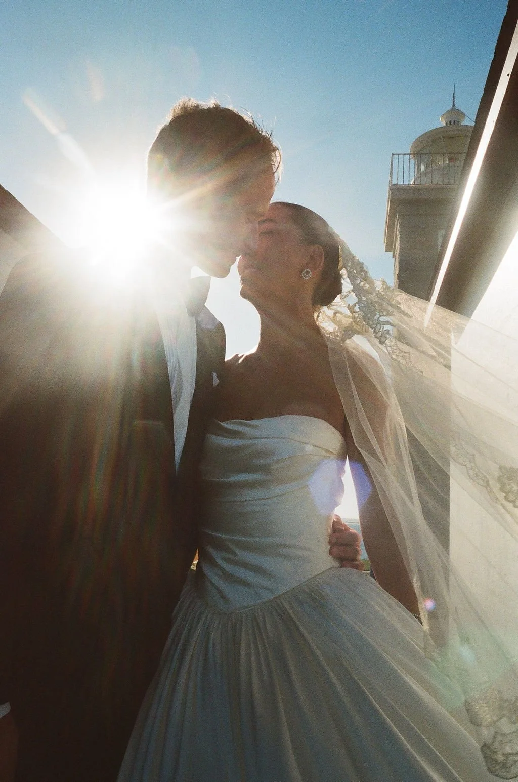 A bride and groom kiss outdoors with the sun shining behind them during sunset, with a building visible in the background.