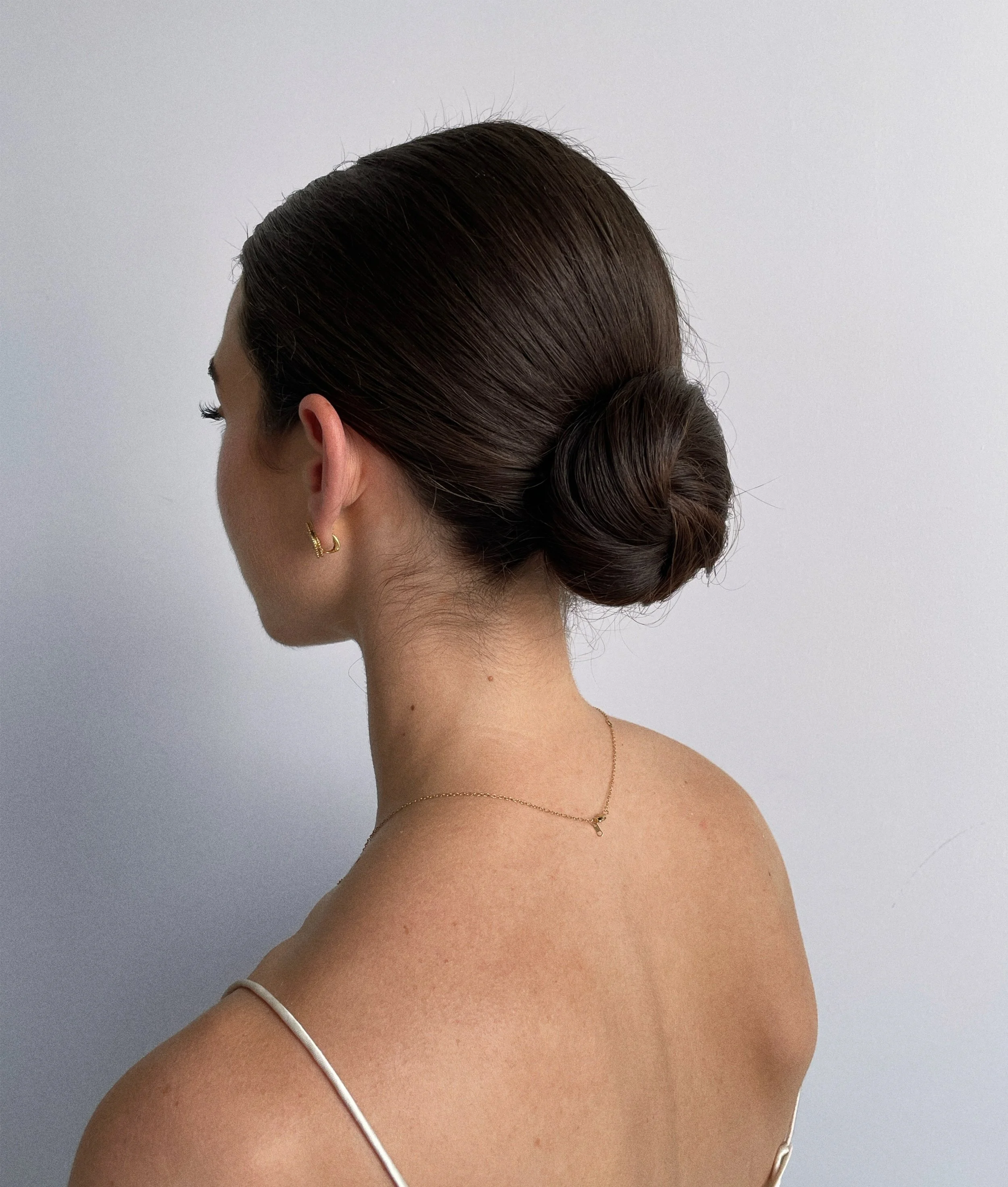 A woman with dark brown hair styled in a low bun, wearing gold earrings and a delicate gold necklace, facing left against a plain light gray background.