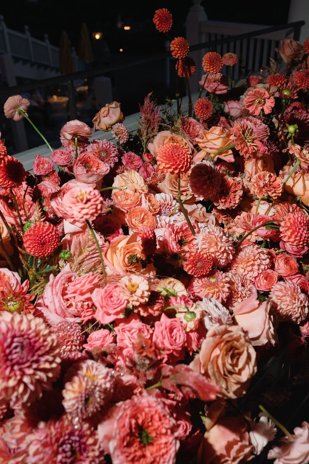 A large collection of pink and peach flowers, including roses, dahlias, and other blooms, arranged outdoors at night.