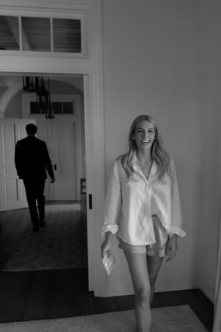 A smiling young woman with long blonde hair, wearing a loose white shirt and shorts, standing in a doorway. A man in a suit is walking away in the background inside a home.