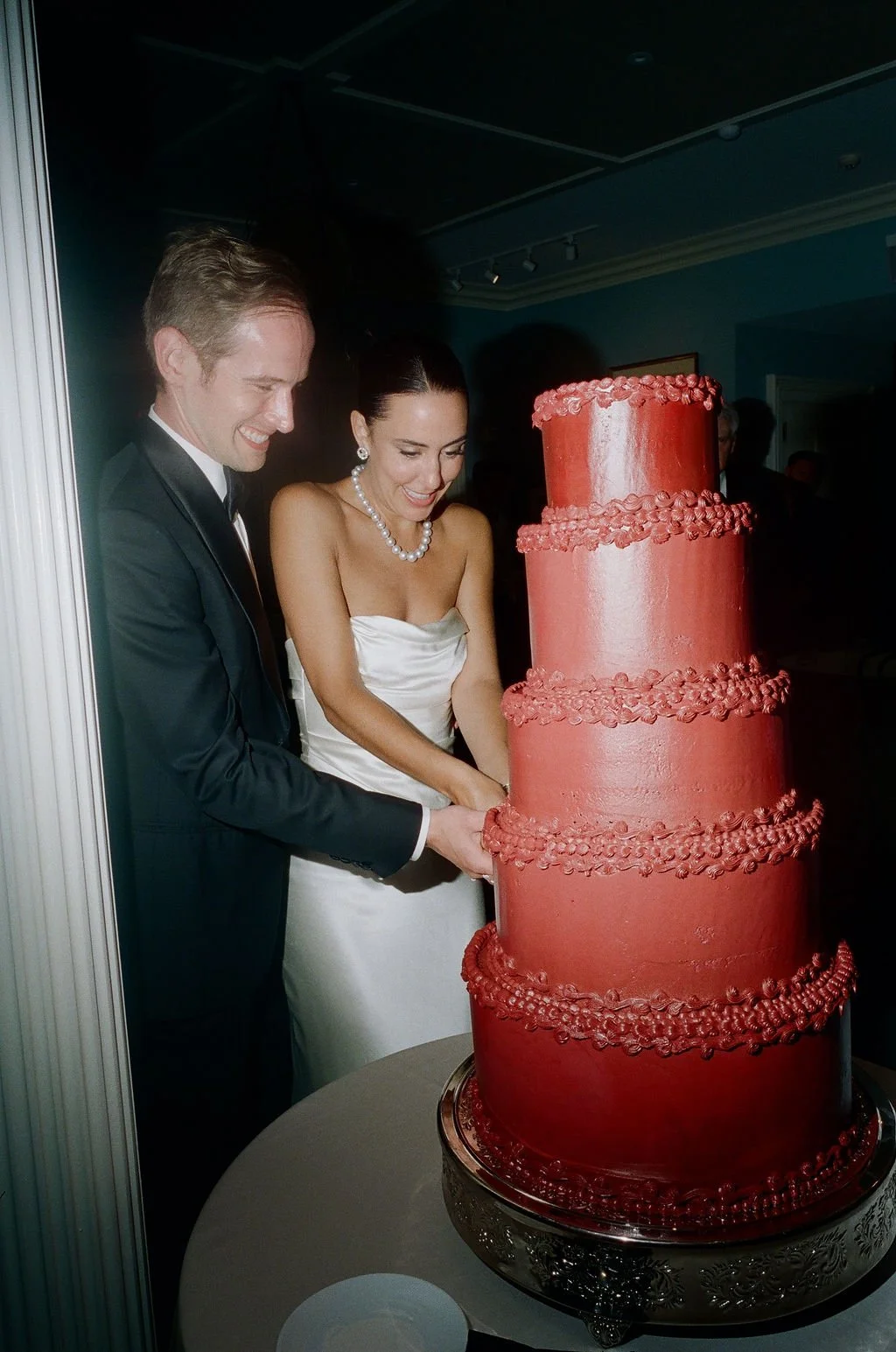 A wedding couple, a man in a tuxedo and a woman in a strapless white dress with pearl jewelry, cutting a large, multi-tiered red wedding cake at their reception.
