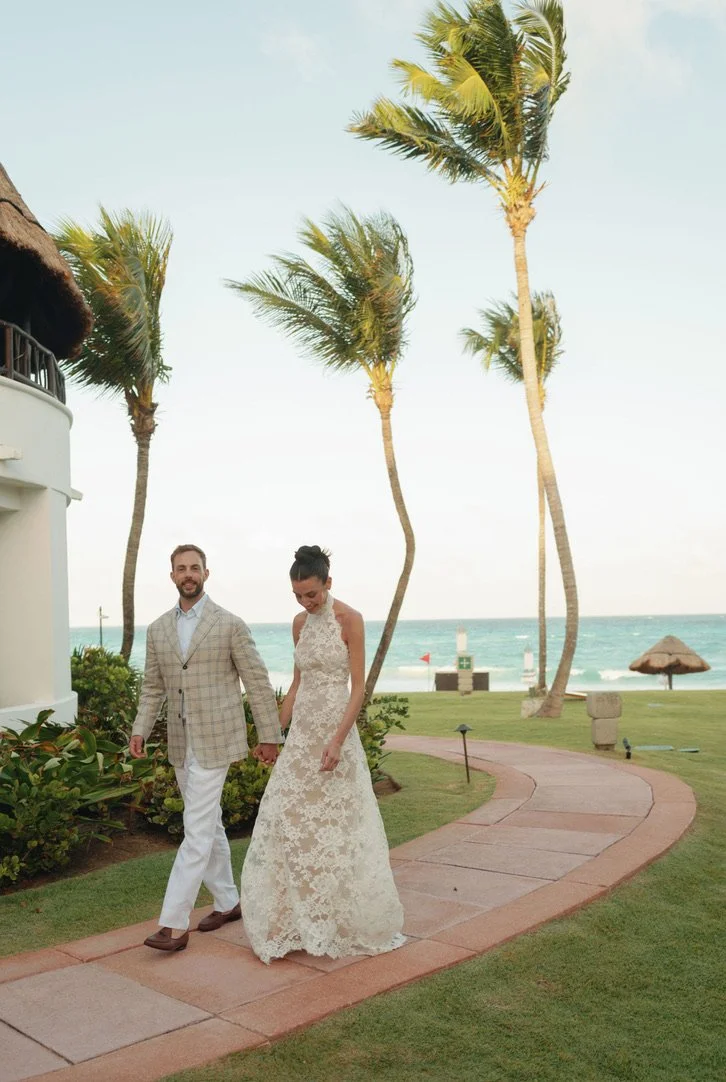 A couple walking hand in hand on a seaside path, with palm trees, ocean, and a thatched hut in the background.