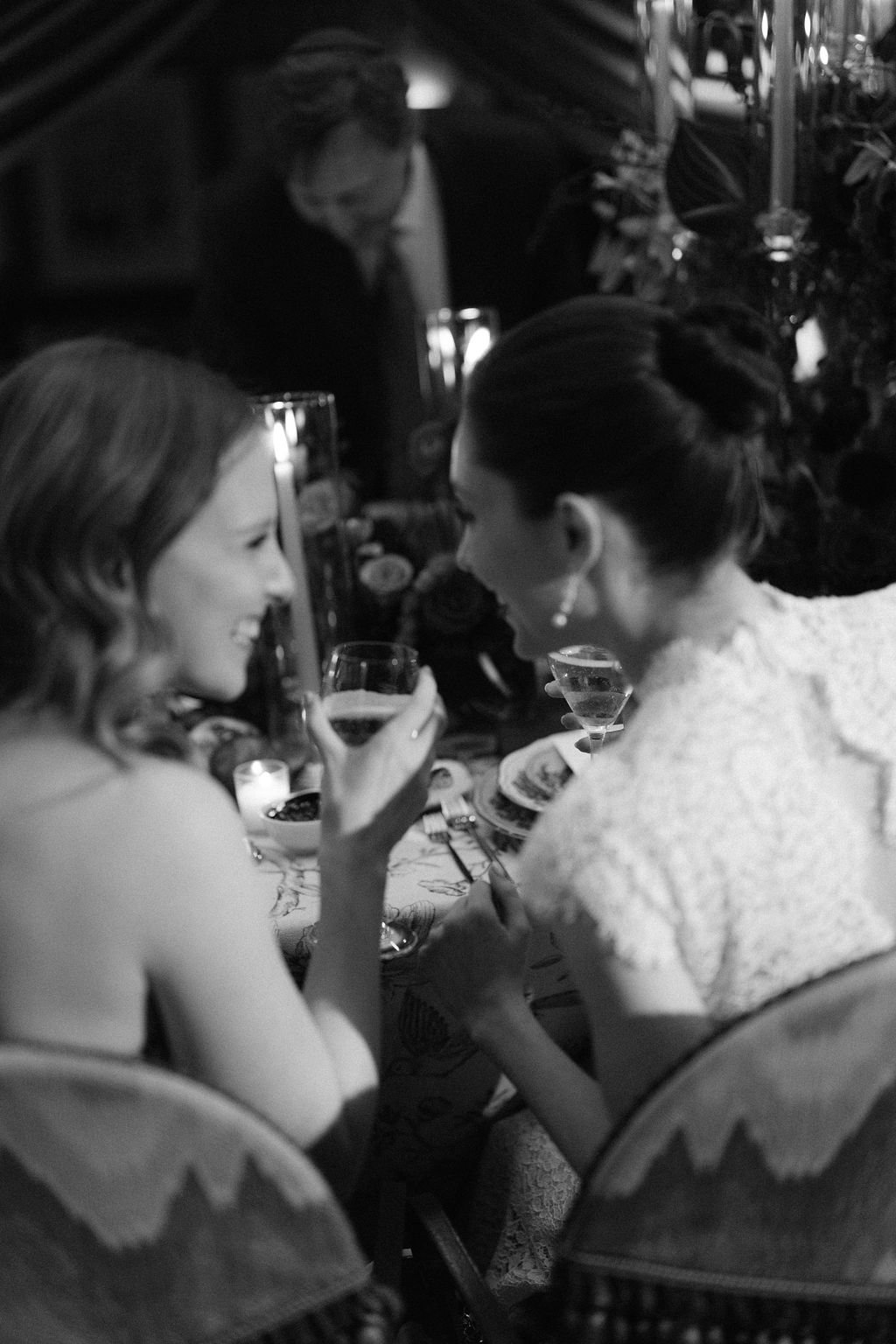 Two women at a dinner table, smiling and holding drinks, in an elegantly decorated setting with candles and floral arrangements. Other people are in the background.
