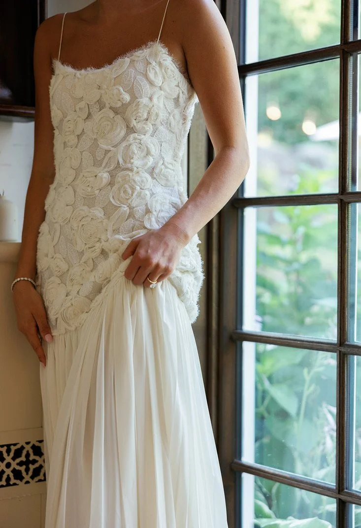 A woman in a white wedding dress with floral appliqué details, standing near a window.