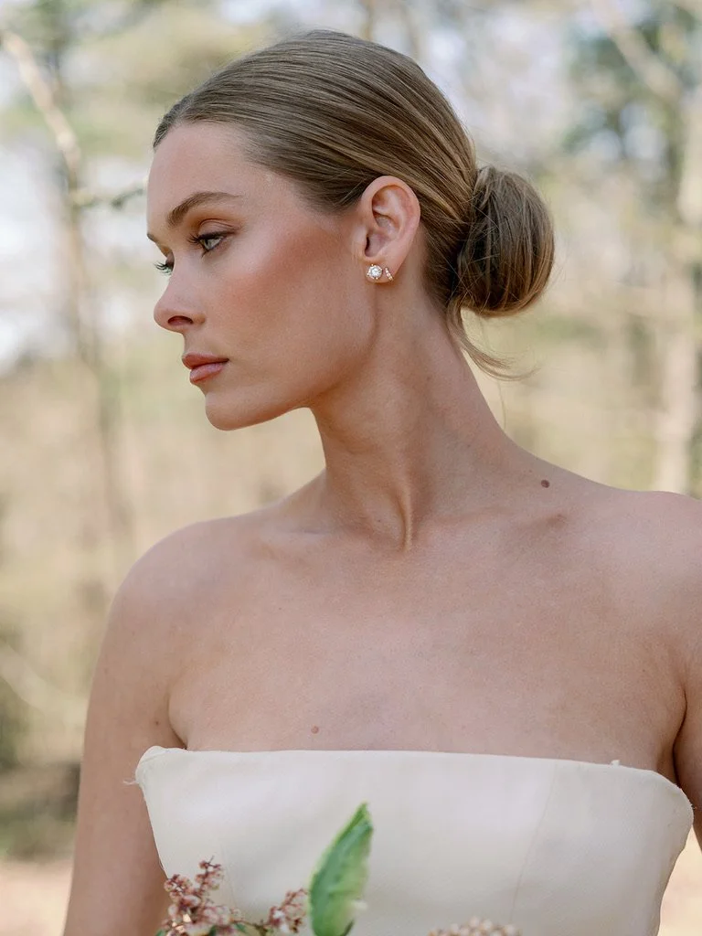 Close-up side profile of a woman with elegant makeup, pearl earrings, and a bun hairstyle, wearing a strapless white dress, outdoors with a blurred background of trees.