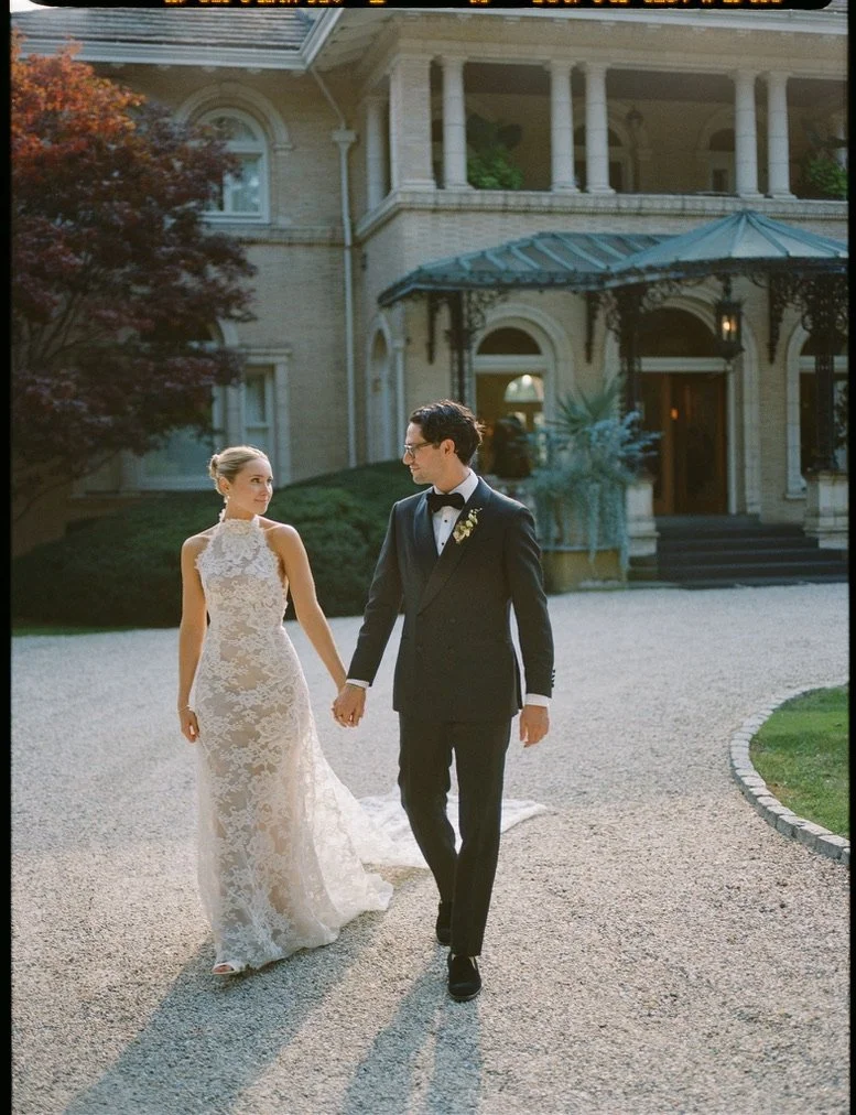 A bride and groom holding hands and walking outside a large, elegant mansion during sunset. The bride wears a lace wedding gown, and the groom is in a black tuxedo with a bow tie.