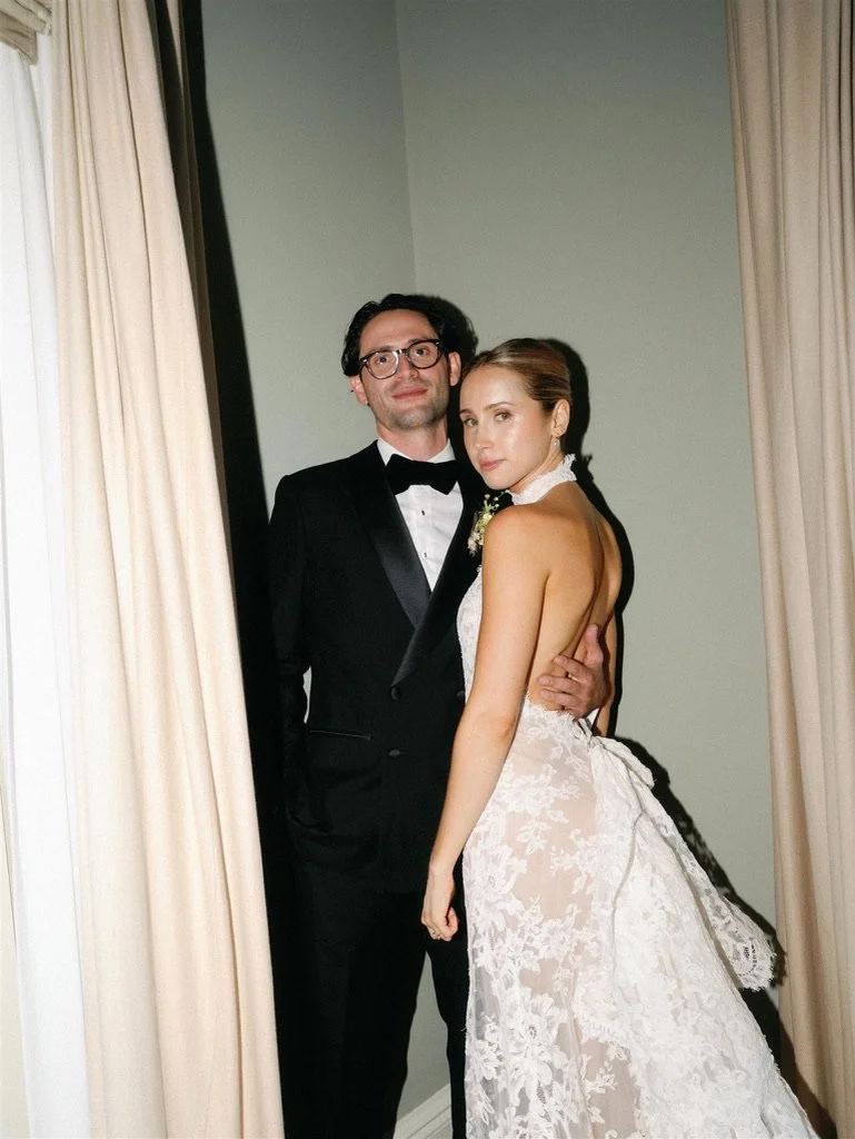 A bride and groom standing close together in wedding attire, inside a room with curtains.