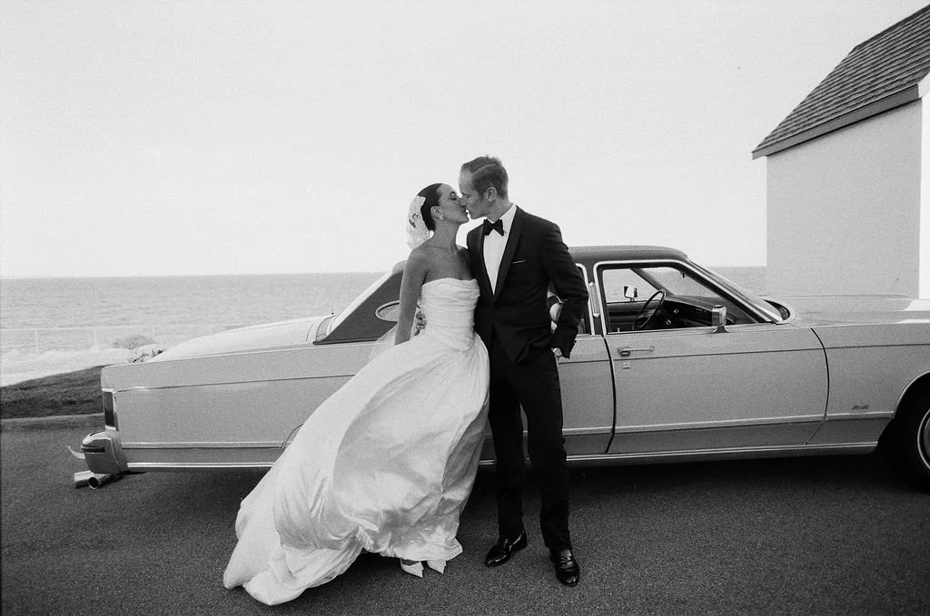 A bride and groom sharing a kiss in front of a vintage car near the ocean, with a building visible to the right.