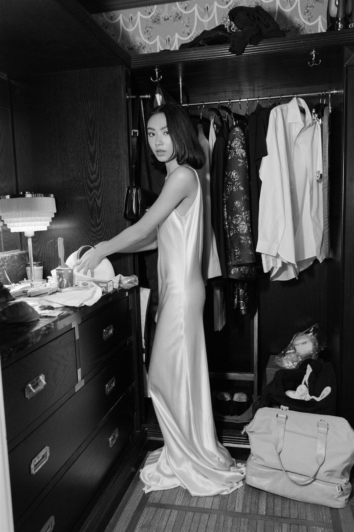 A woman in a silky dress standing in a hotel closet, surrounded by clothes and personal items.