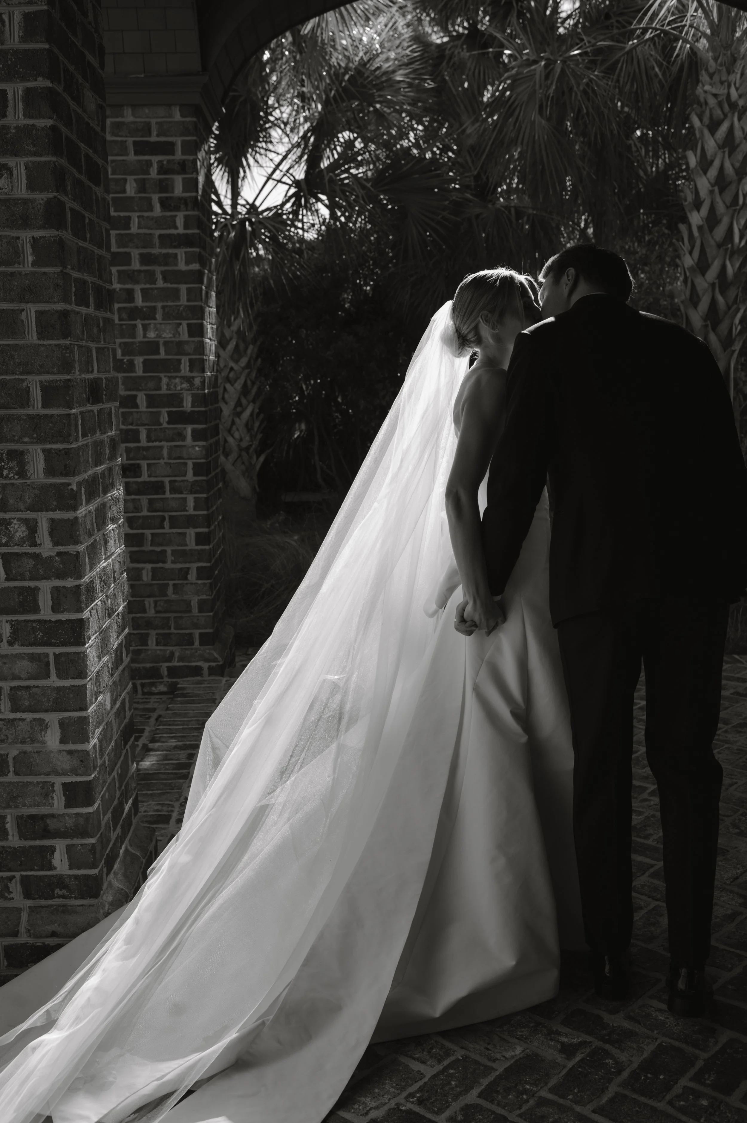 Black and white photo of a wedding couple at sunset, with the bride in a wedding dress and veil, and the groom in a suit, holding hands, standing close together outdoors.