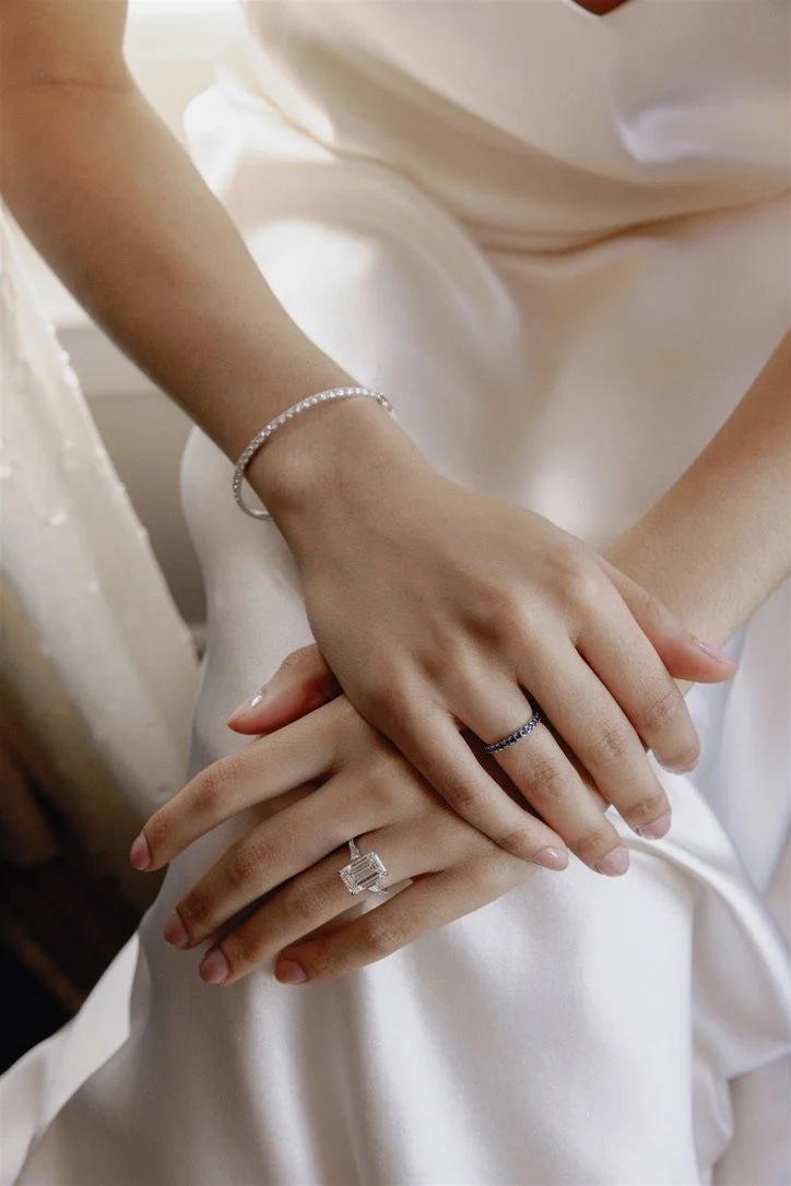Close-up of a woman's hands and wrists resting on her lap, adorned with rings and a bracelet, dressed in a white satin garment.