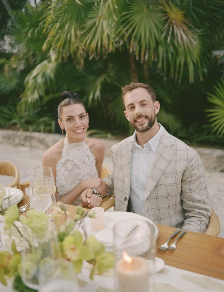 A smiling man in a light-colored plaid blazer and a woman in a white lace dress sitting at an outdoor table surrounded by green tropical plants.