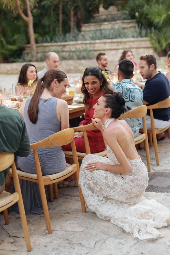 A wedding reception outdoors with guests sitting at a long table, smiling and chatting. A bride in a white lace dress is kneeling on the ground, leaning on a chair, talking to a woman in a red dress.