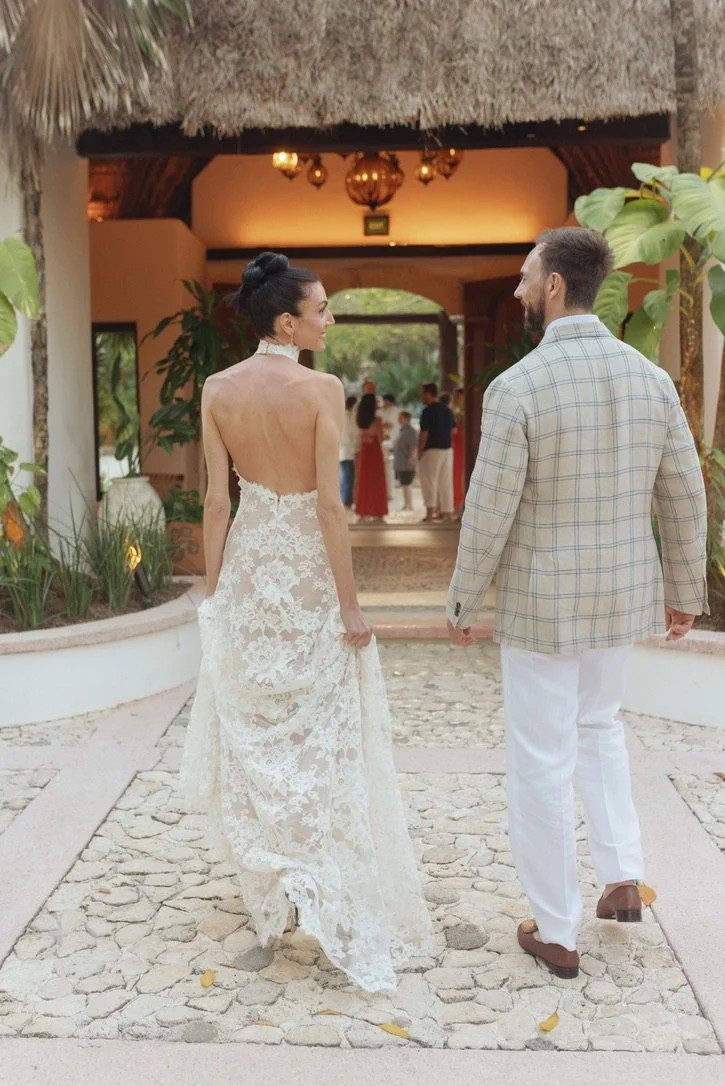 Bride and groom holding hands and walking outdoors with tropical plants and a thatched-roof structure in the background, at a wedding or celebration.