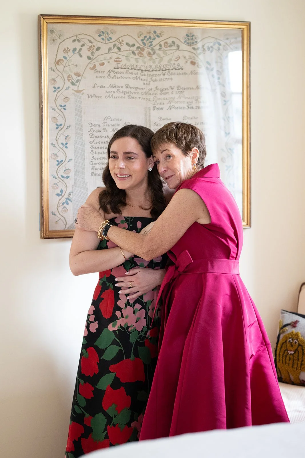 A young woman in a floral dress embracing an older woman in a bright pink dress in a warmly lit room, with a framed embroidered family tree on the wall behind them.