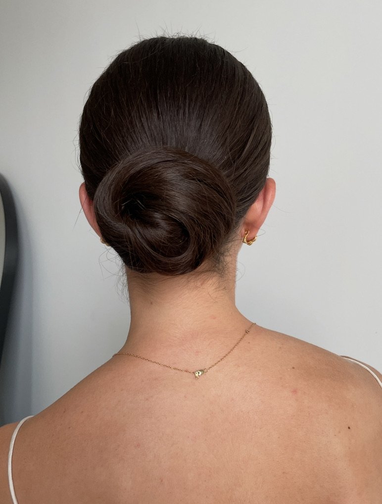 Back view of a woman with dark brown hair styled in a sleek bun, wearing gold earrings and a delicate gold necklace, with a light gray background.