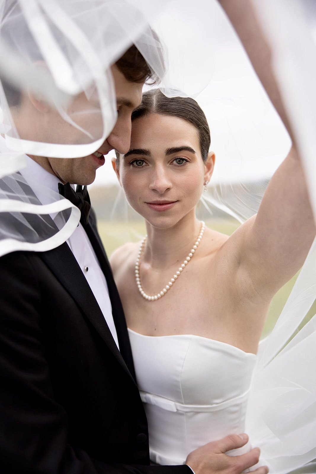 A bride and groom in wedding attire, standing close together outdoors. The bride is looking at the camera, wearing a strapless white dress and pearl necklace, with a sheer veil over her head. The groom, in a black tuxedo with a bow tie, is leaning to