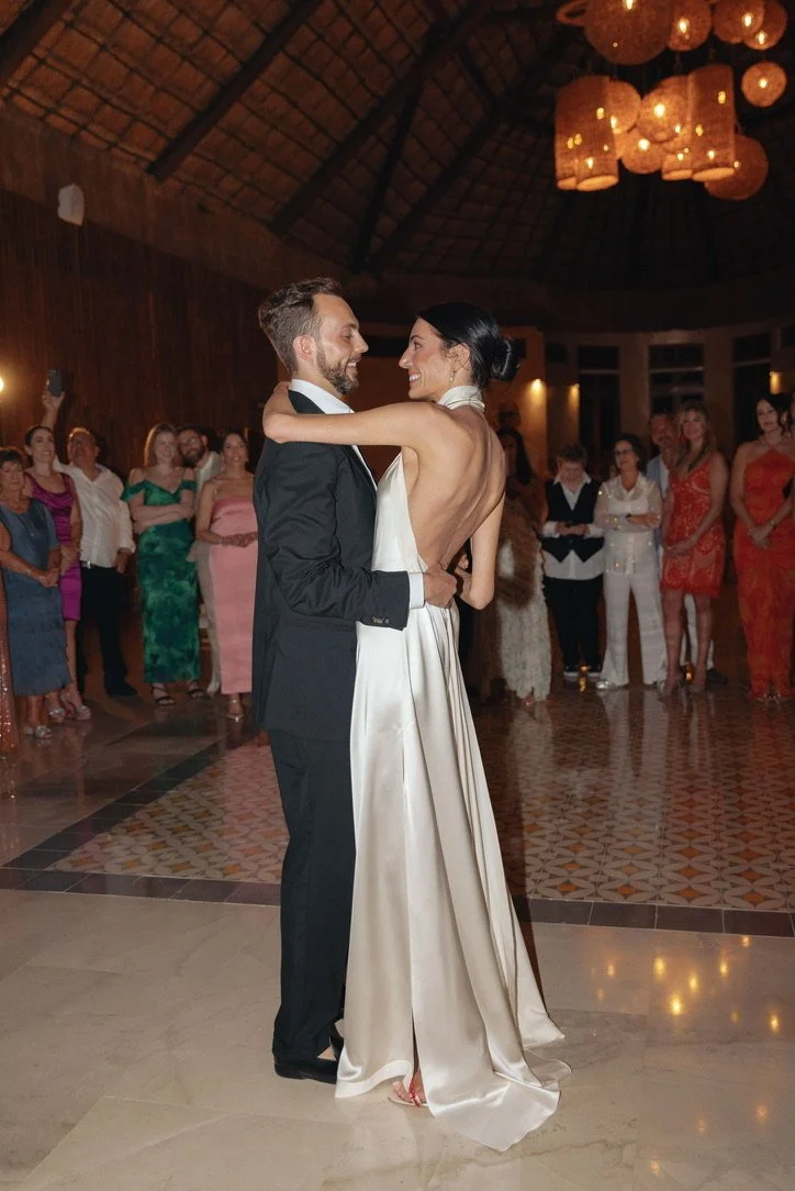A bride and groom are dancing and smiling at their wedding reception wearing Danielle Frankel with guests watching in the background.