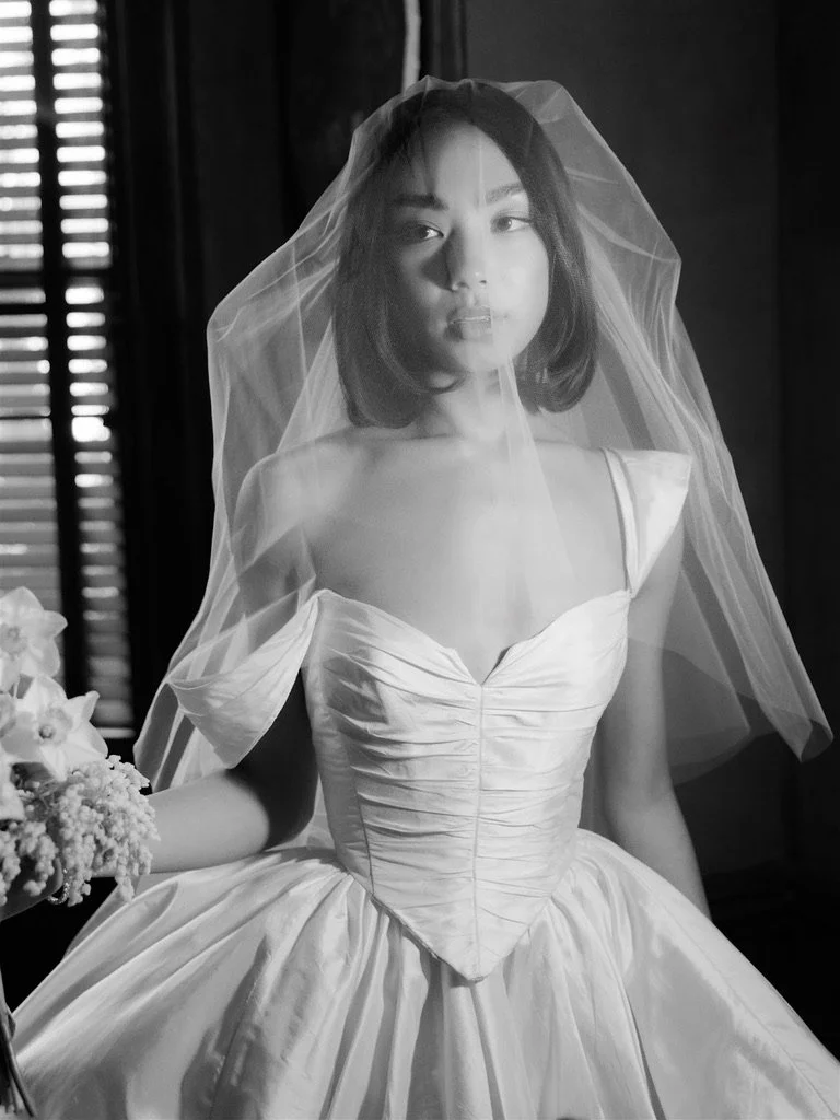 Black and white photo of a woman in a wedding dress with a veil, standing indoors near a window.
