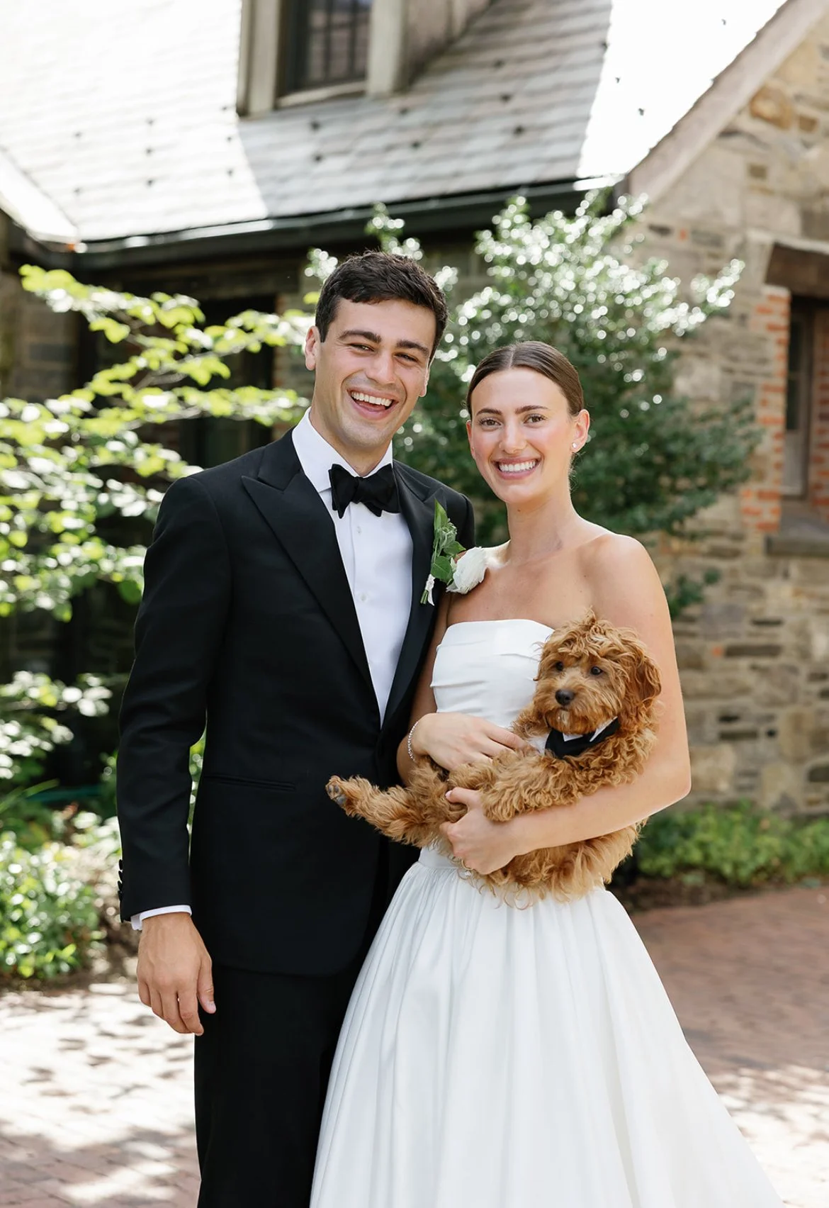 A smiling bride and groom dressed in wedding attire, holding a small brown puppy wearing a tuxedo, outdoors in front of a stone building and greenery.