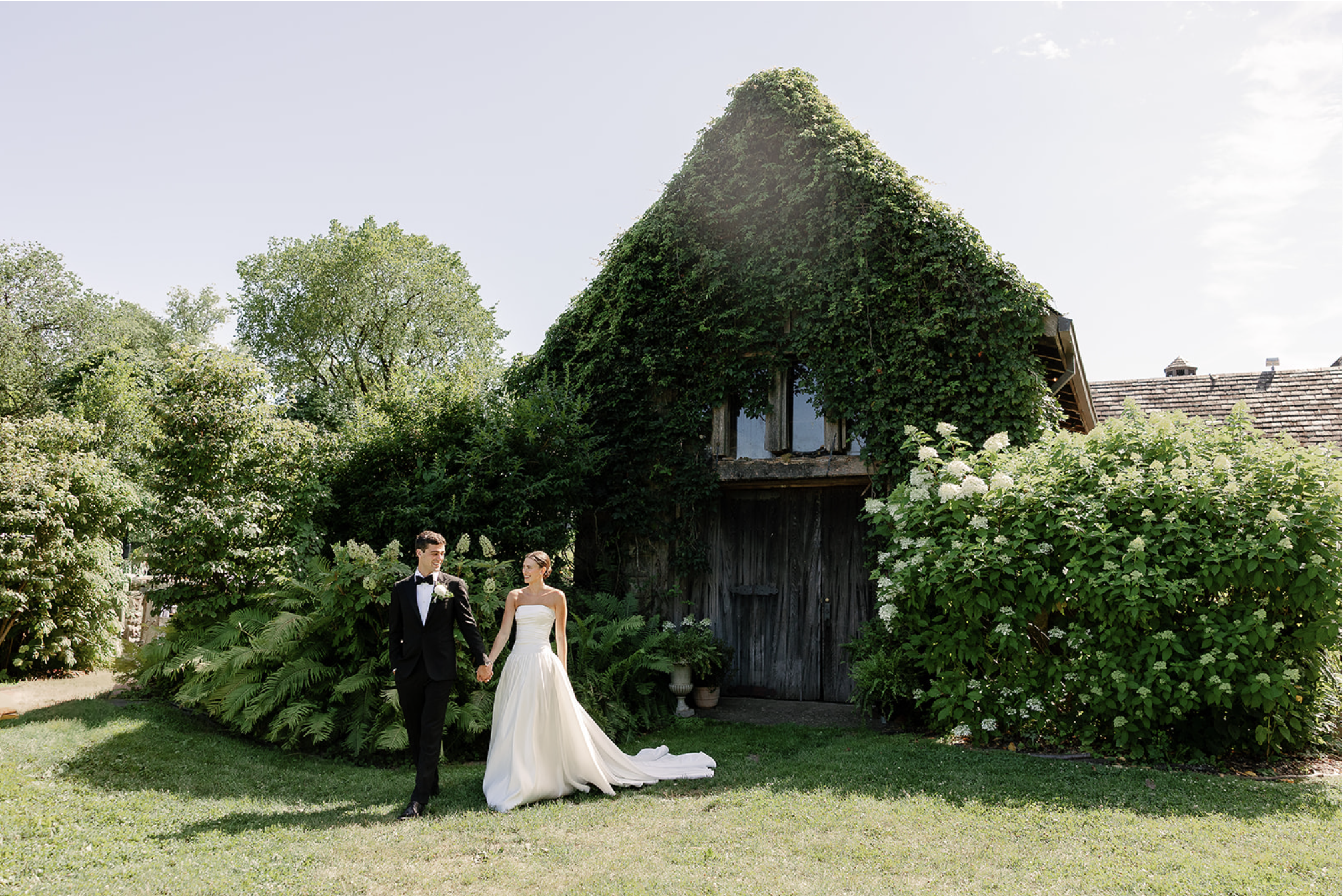 A bride and groom holding hands, walking in a garden with lush greenery and an ivy-covered rustic barn in the background.