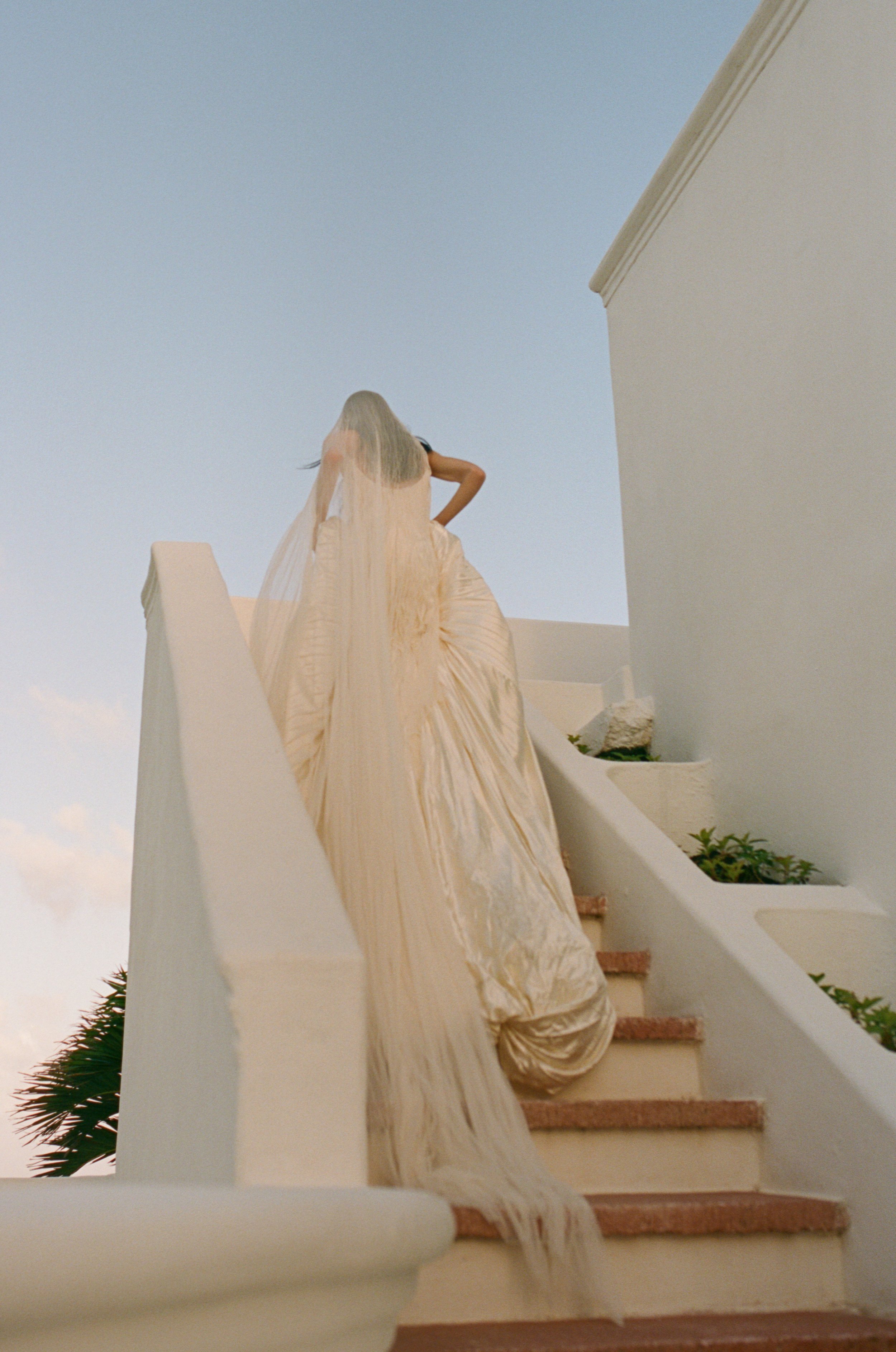 A woman wearing a flowing, cream-colored dress with a long train, standing on a white staircase outdoors during sunset, with a palm tree visible in the background.