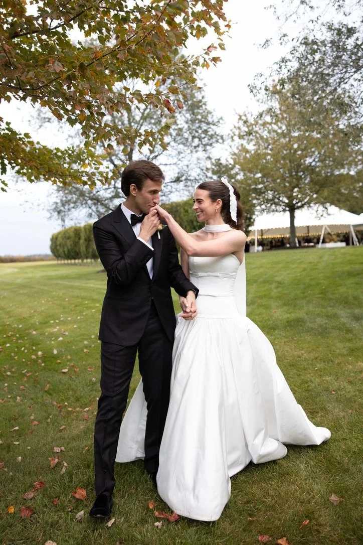 A bride and groom in wedding attire sharing a tender moment outdoors on a grassy field, with trees and a tent visible in the background.