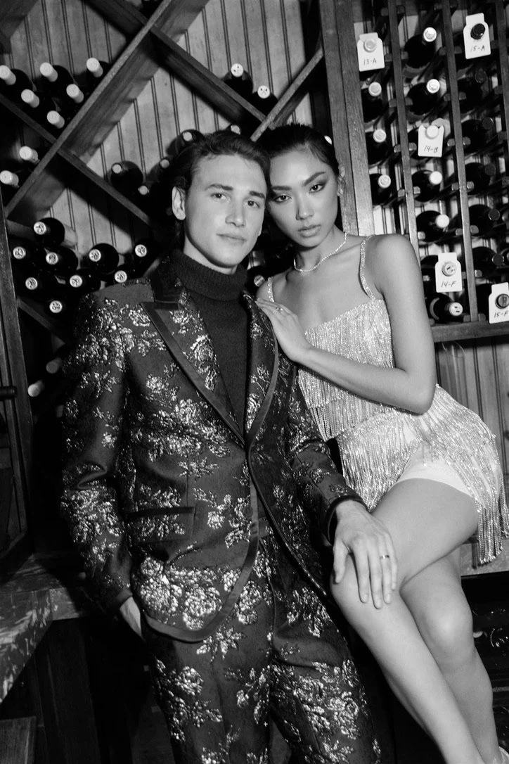 A black and white photo of a man and woman posing together in a wine cellar with wine bottles on wooden shelves behind them. The man is wearing a patterned suit and turtleneck, and the woman is in a sparkly dress with fringe details.