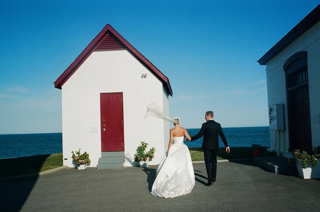A bride and groom holding hands walking near a small white chapel with a red door, by the ocean on a clear day.