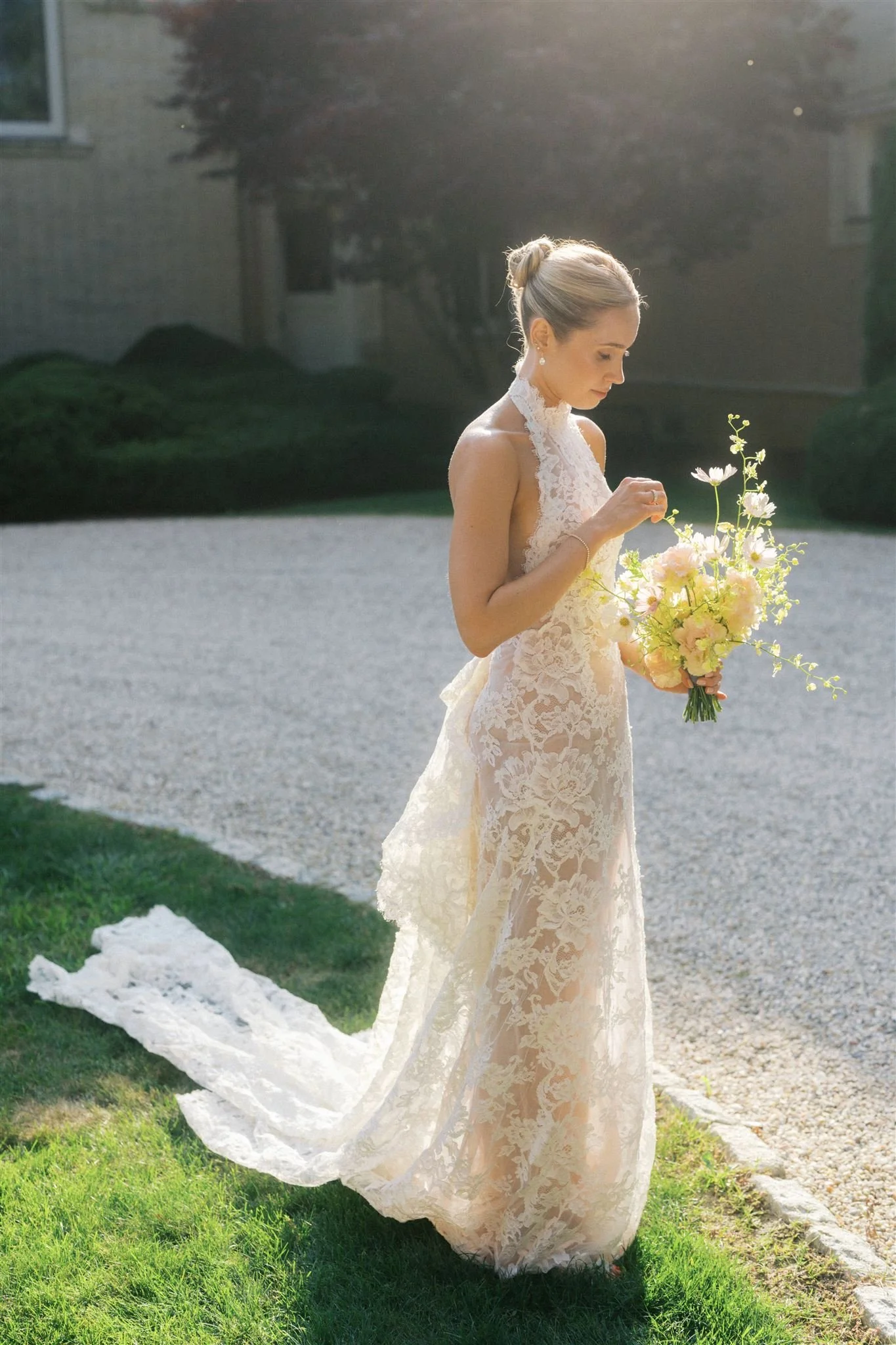 A bride in a lace wedding gown holding a bouquet of pink and yellow flowers, standing on a grassy area with sunlight shining behind her.