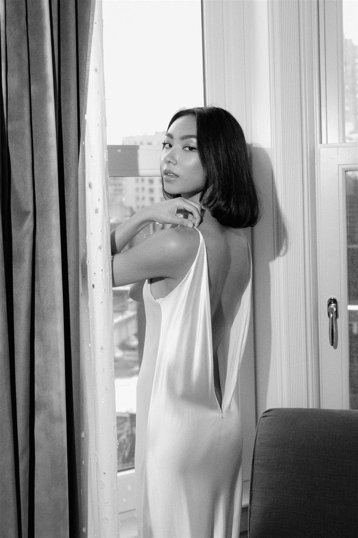 A woman with shoulder-length hair standing by a window, wearing a backless satin dress, looking at the camera.