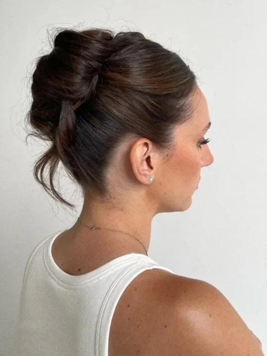 Side profile of a woman with styled brown hair in an updo, wearing a white sleeveless top and small earrings, against a plain light background.