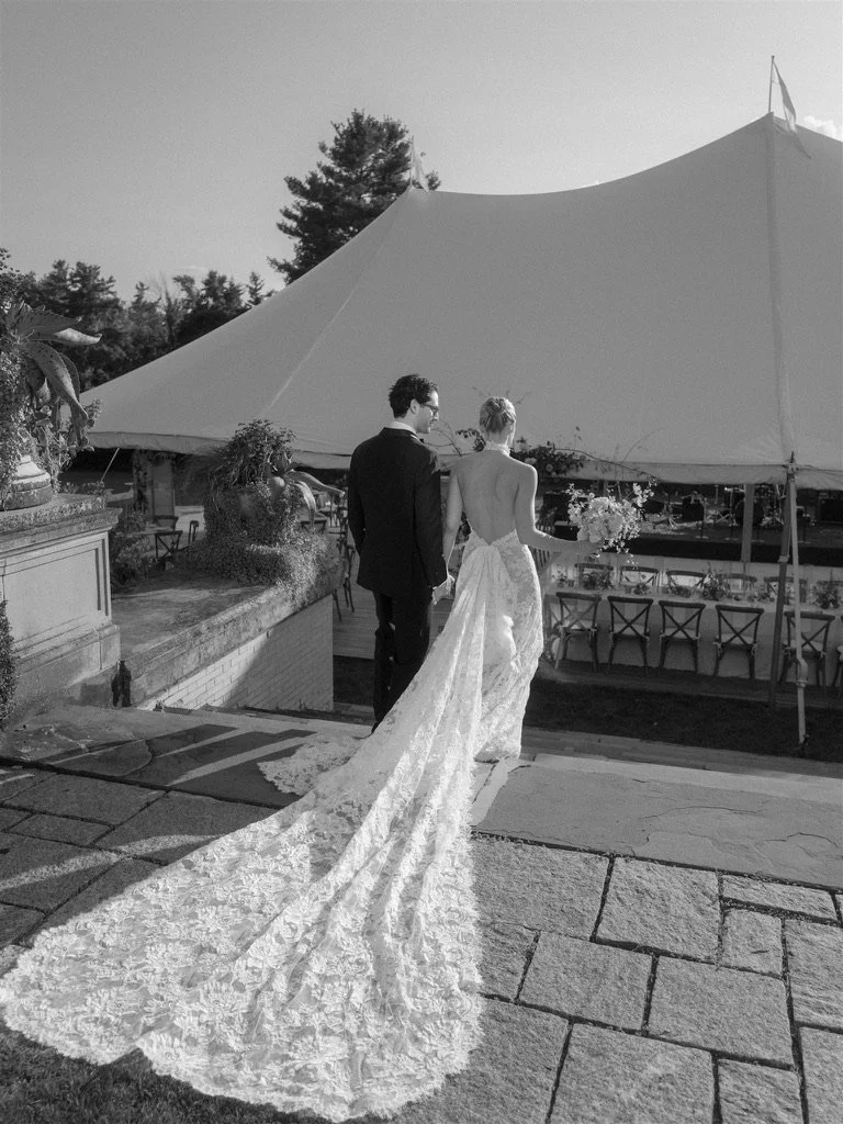 Bride and groom walking on stone path at outdoor wedding reception; bride in a long lace train dress, groom in a dark suit; large tent and outdoor seating in background.