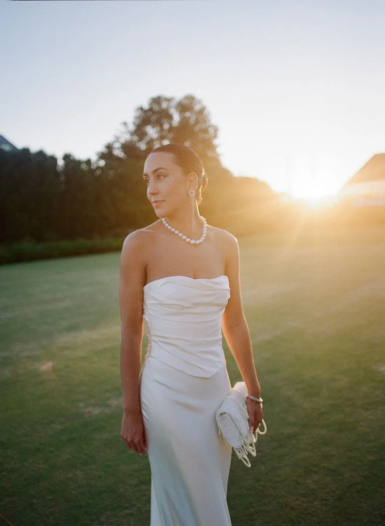 A woman in a white strapless dress and pearl necklace standing outdoors during sunset, holding a white clutch purse.