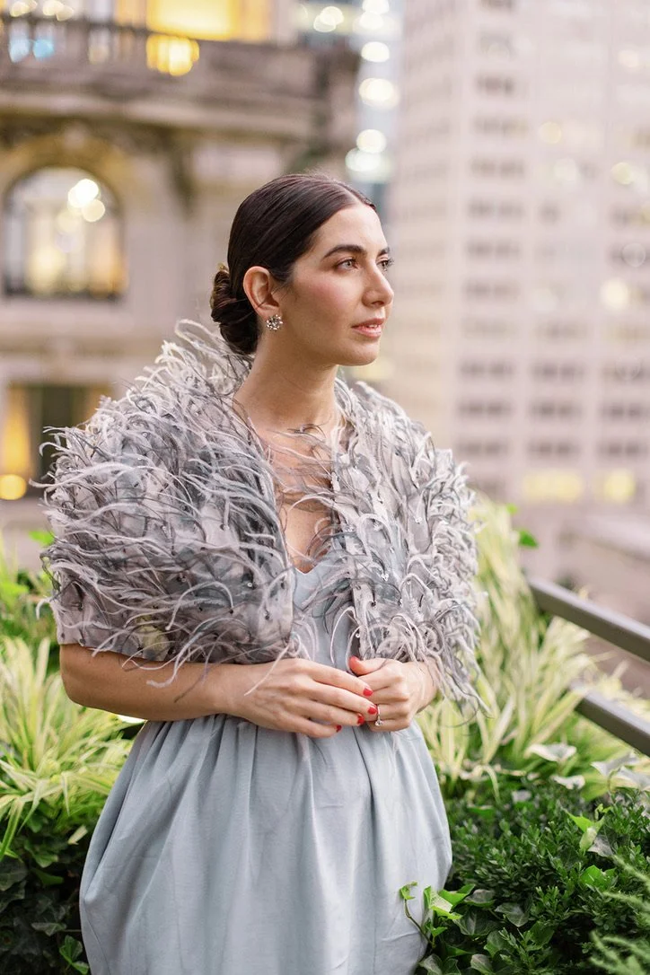 A woman with dark hair styled in an elegant updo, wearing a gray dress and a feathered shawl, standing outdoors in an urban setting with buildings and greenery in the background.