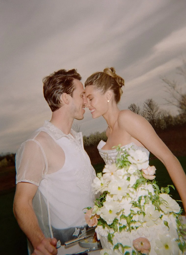 A smiling couple, dressed in wedding attire, leaning their foreheads and noses together outdoors during daytime, with trees and cloudy sky in the background.